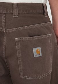 Pruunid tööpüksid, mis on valmistatud vastupidavast kangast, tagaküljel on tasku halli ja oranži Carhartt logo ning õmblused on märgatavad kontrastse värviga.