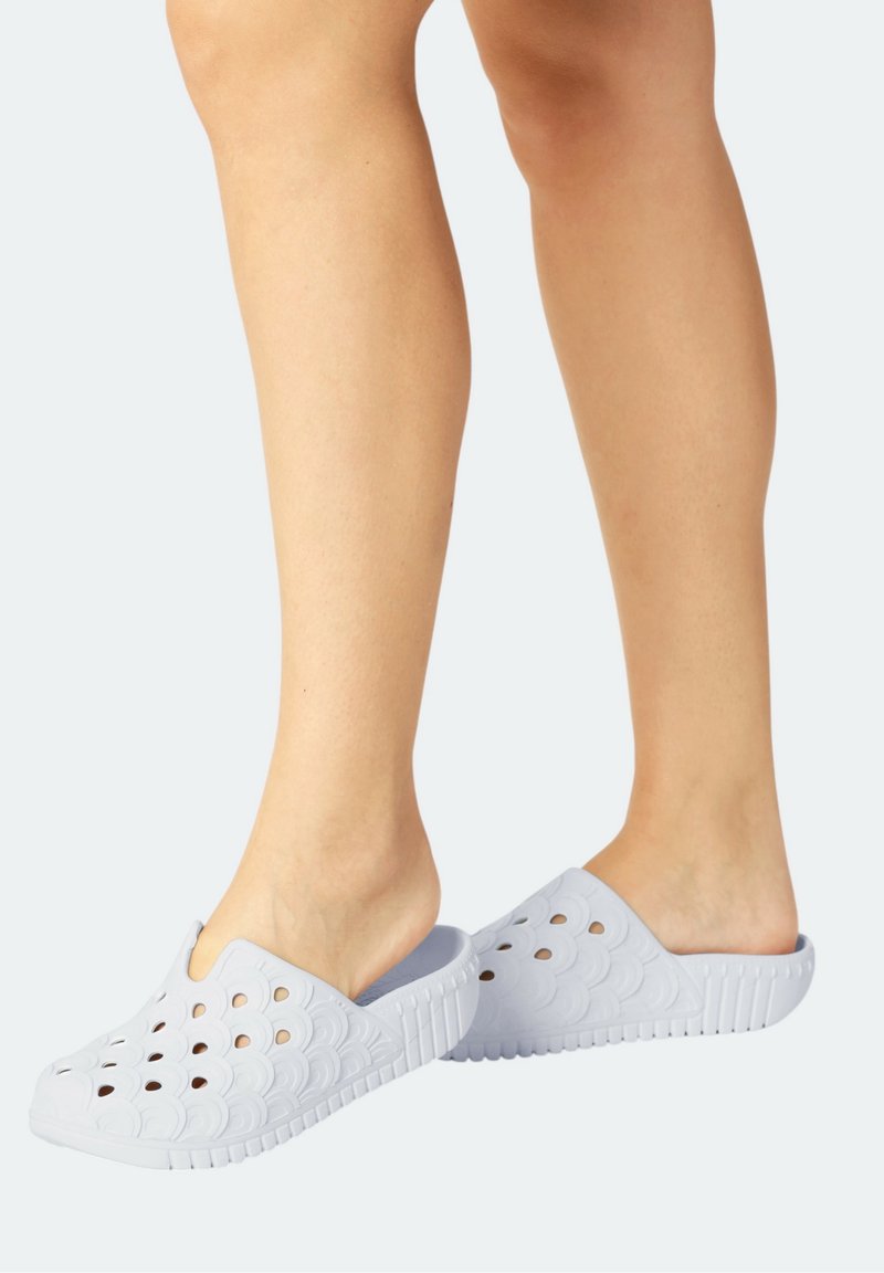 Jambes portant des chaussures blanches à enfiler avec des trous perforés et une semelle texturée, debout sur un fond gris pâle.