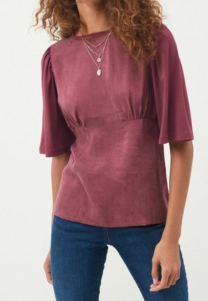 Kobieta w fioletowej bluzce z bufkami do łokcia i warstwowymi srebrnymi naszyjnikami, w połączeniu z niebieskimi dżinsami.