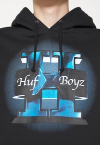 Sudadera negra con un gran diseño gráfico que presenta acentos azules, el texto "Huf X Boyz" y un número "11" en un estilo en capas.