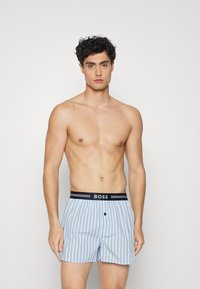 Boxers à rayures bleues avec une taille noire arborant le nom de la marque "BOSS" en blanc. Fermeture à bouton sur le devant et tissu léger.