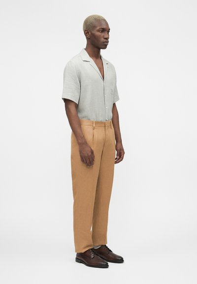 Άνδρας με κοντά ξανθά μαλλιά, φορούντας ένα ανοιχτό γκρι κοντομάνικο πουκάμισο με κουμπιά, καφέ παντελόνια και σκούρα καφέ δερμάτινα παπούτσια, όρθιος πλευρικά.