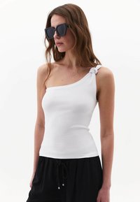 Top sin mangas blanco de un solo hombro con un detalle de nudo en la tira, combinado con pantalones negros con cordón. Textura de tela suave con una silueta ajustada.