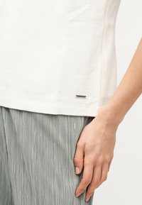 Άτομο που φοράει λευκό υφασμάτινο κοντομάνικο μπλουζάκι με μικρή ετικέτα λογοτύπου και ανοιχτό γκρι πλισέ παντελόνι, με το χέρι ακουμπισμένο στο πλάι.