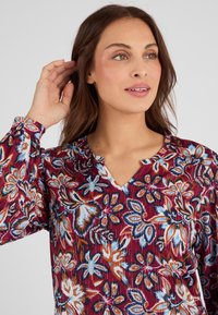 Blusa floreale con uno sfondo bordeaux intenso, arricchita da intricati motivi blu, arancioni e bianchi, e un design a V con leggere pieghe.