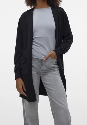 Personne portant un haut bleu clair, un long cardigan noir ouvert et un jean gris clair avec une main dans la poche sur un fond uni.