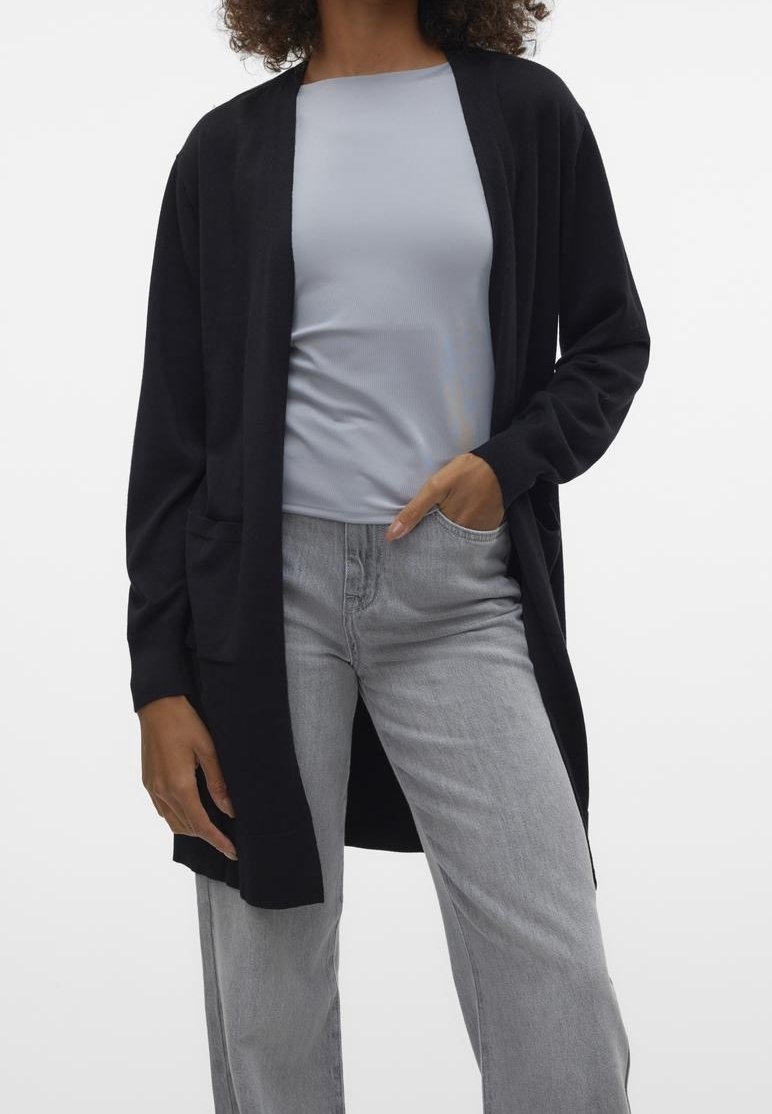 Personne portant un haut bleu clair, un long cardigan noir ouvert et un jean gris clair avec une main dans la poche sur un fond uni.