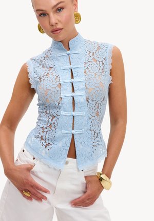 Woman wearing sleeveless light blue lace top with frog closures, white pants, large gold spiral earrings, and gold bracelet and ring.