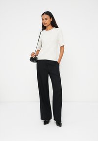 Gai bēšas īsu piedurkņu blūze ar apaļu kakla izgriezumu, apvienota ar melniem svītrainiem platām biksem un mazu melnu somiņu.