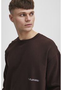 Brązowa bluza z okrągłym dekoltem, wykonana z miękkiego materiału. Posiada białe logo tekstowe po lewej stronie oraz luźny krój.