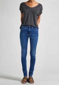 Camiseta gris de cuello en V combinada con jeans skinny azules. Las sandalias tienen múltiples tiras y tachuelas. En general, un atuendo casual y ajustado.