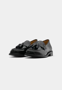 Black patent leather loafers with a smooth finish, featuring decorative tassels and a low, solid heel. Structured shape with visible stitching.