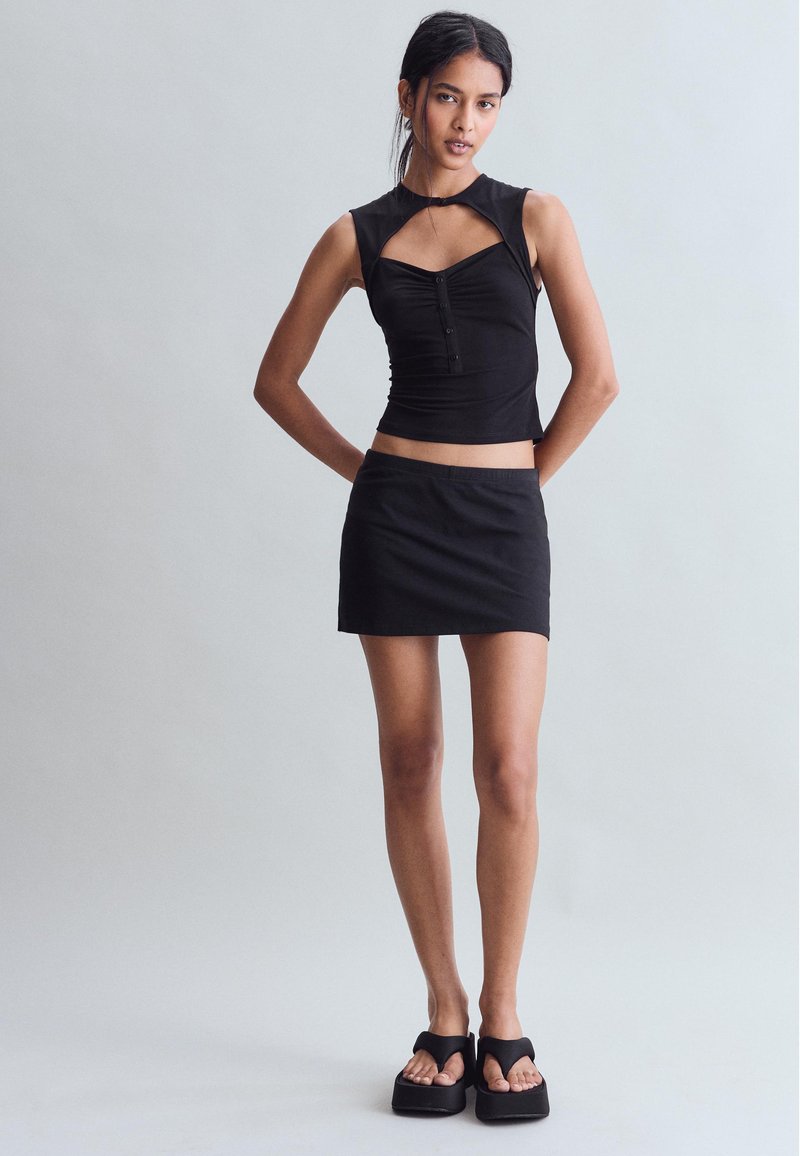 Femme debout portant un haut noir sans manches avec découpes, une mini-jupe noire et des sandales compensées noires sur un fond clair uniforme.