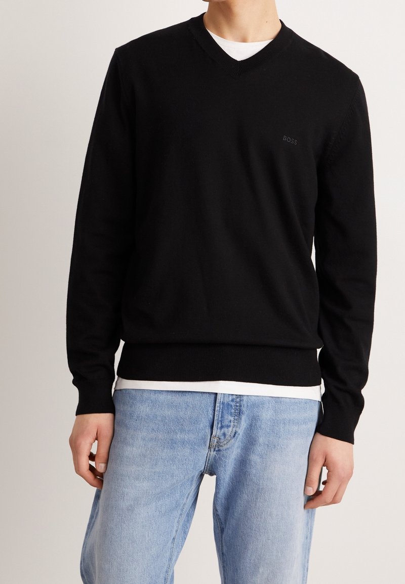 Homme portant un pull noir à col en V par-dessus une chemise blanche avec un jean bleu clair, debout devant un fond clair uni.
