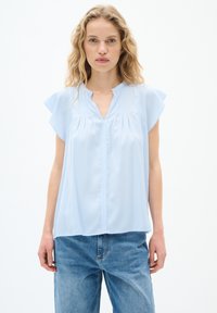 Blusa blu chiaro con maniche a sbuffo corte, scollatura a V, pieghe sul davanti, tessuto morbido e taglio dritto; abbinata a jeans blu.