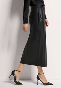 Lange schwarze Lederrock mit glatter Textur, hoher Taille und dezenten Falten. Kombiniert mit eleganten schwarzen Stilettos.