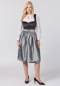 Traditionelles Dirndl mit dunkelblauem Mieder, weißer Bluse mit Lochmuster und glänzendem silbernen Rock mit Spitzenbesatz. Kombiniert mit schwarzen Absätzen.