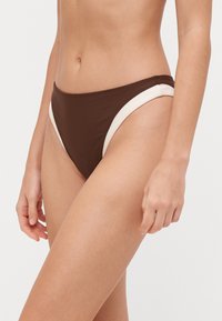 Brūns bikini apakšda ar krēmkrāsas sānu paneļiem, ar gludu auduma tekstūru un vidēji augstu jostasvietu. Minimālistisks dizains bez rotājumiem.
