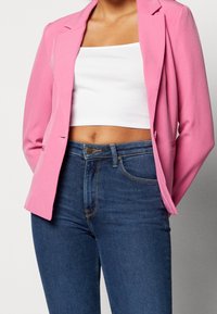 Kobieta w jaskraworóżowej marynarce, białym skróconym topie i ciemnoniebieskich dżinsach z wysokim stanem, stojąca na jednolitym tle.