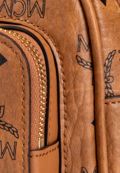Brown leather bag with gold zipper and black printed MCM logo and laurel pattern, featuring detailed stitching and a leather strap.