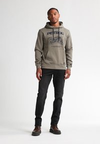 Sudadera verde oliva con un estampado gráfico, bolsillo canguro, capucha con cordones; combinada con jeans negros y zapatos marrones con cordones.