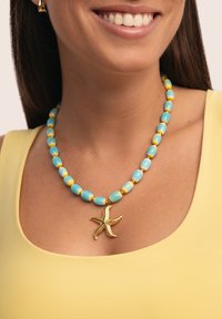 Un collar de cuentas azules y amarillas presenta un elaborado colgante en forma de estrella de mar dorada, descansando sobre una blusa amarilla con escote redondeado.
