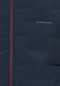 Τμήμα σε μανδί στρατιωτικού μπλε με κάθετο κόκκινο και μαύρο φερμουάρ στα αριστερά και λευκό λογότυπο "Jack & Jones" στη δεξιά πλευρά.