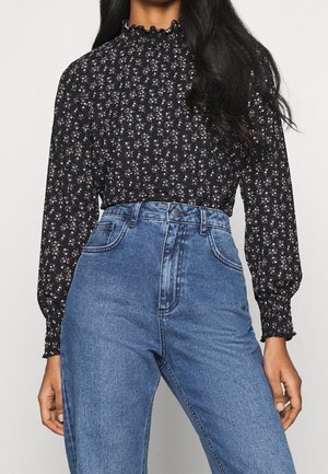 Vrouw die een zwarte, lange mouwen bloemenblouse draagt met geplooide manchetten en een hooggetailleerde blauwe spijkerbroek, staand met ontspannen armen.