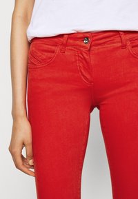 Pantalones vaqueros de mezclilla rojos con un ajuste Slim, diseño de cinco bolsillos y cierre de botón. Textura suave con detalles de costura a lo largo de los bolsillos.