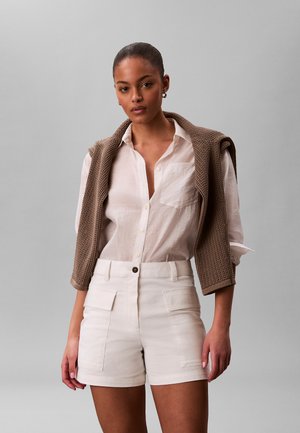 Mujer que lleva una camisa blanca con botones y pantalones cortos blancos, con un suéter marrón de punto apoyado sobre los hombros, contra un fondo gris liso.