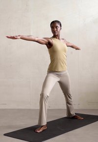 Una canotta atletica beige e pantaloni yoga color crema su una persona in posizione guerriero su un tappetino nero. Tessuto morbido con un design aderente. Sfondo neutro.