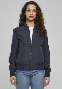 Geacă bomber bleumarin din material ușor, cu fermoar în față, mansete striate și talie ajustată. Purtată peste o cămașă albă.