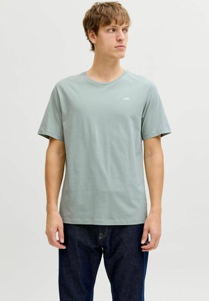 Joven con camiseta de manga corta de color verde claro con pequeño logo blanco "J.J.R.", combinada con vaqueros azul oscuro, de pie frente a un fondo liso.