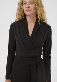 Juoda apvyniojimo suknelė su ilgomis rankovėmis, V formos iškirpte ir raukšlėta detalizacija krūtinėje. Suknelė turi surištą liemenį ir lygų, švelnų audinio tekstūrą.