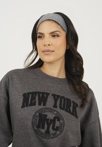 Sweatshirt gris avec un motif noir "NEW YORK" et un logo circulaire au centre. Porté avec un bandeau gris assorti. Texture lisse, coupe décontractée.