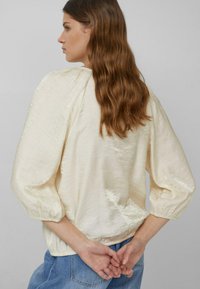 Kobieta z długimi falującymi brązowymi włosami, ubrana w błyszczącą kremową bluzkę z bufkami i niebieskie dżinsy, stoi ze złączonymi rękami za plecami.