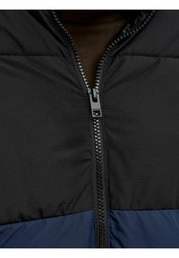 Schwarze und marineblaue gesteppte Jacke mit hohem Kragen, durchgehendem Frontreißverschluss und sichtbaren Nähdetails auf dem Stoff.