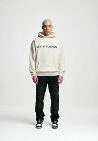 Felpa crema con grande testo nero "2Y STUDIOS", tasca a marsupio frontale, abbinata a jeans neri e sneaker bianco e neri.