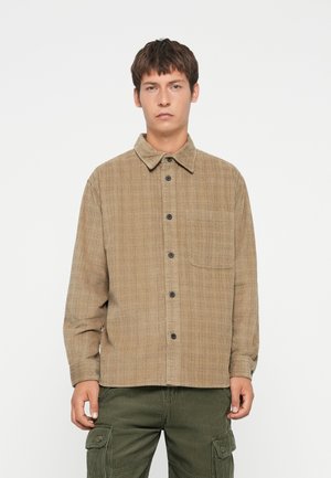 Brown plaid button-up shirt with a collar, chest pocket, and black buttons; made of textured fabric, paired with green cargo pants.