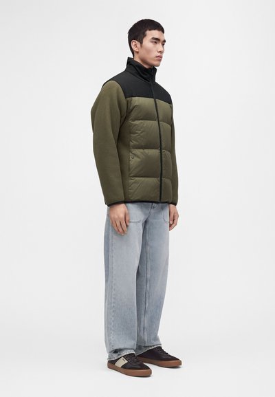Jeune homme debout portant une doudoune vert olive et noire, un jean large bleu clair et des baskets noires avec des semelles marron.
