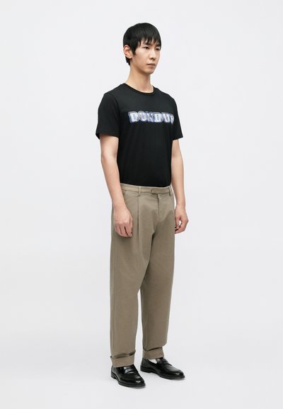 Μαύρο μπλουζάκι με γραφικό σχέδιο "DONDUP". Μπεζ παντελόνι, ελαφρώς φαρδύ, με ρεβέρ στους αστραγάλους. Μαύρα παπούτσια, απλός σχεδιασμός. Ουδέτερο φόντο.