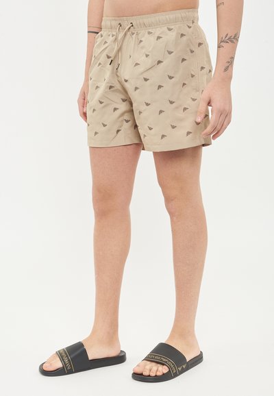 Pantaloni da bagno beige con coulisse in vita, caratterizzati da un motivo a triangoli ripetuto di colore marrone scuro. Abbinati a sandali neri a scivolo.