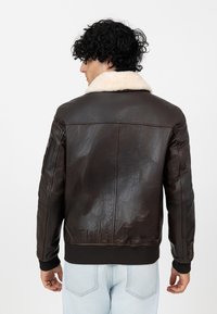 Bruine leren jack met een witte schapenvachtkraag, geribbelde manchet en tailleband, en een ritssluiting zak op de linkerarm.