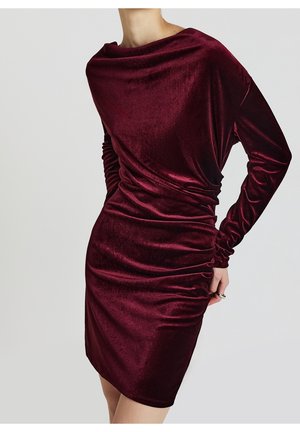 Vrouw draagt een nauwsluitende, lange mouwen fluwelen jurk in donkerrood met geplooide zijdetails, staand tegen een effen lichtgrijze achtergrond.