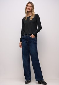 Schwarzes langärmeliges Oberteil kombiniert mit weiten, dunkelblauen Jeans. Die Jeans haben Fronttaschen und einen rohen Saum, ergänzt durch schwarze Stiefeletten.