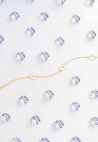 Bracelet en chaîne dorée avec des accents hexagonaux, entouré de pierres claires comme des cristaux taillés, sur un fond blanc lisse.