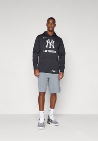 Juodas gobtuvas su "Yankees" logotipu, pilkos šortai, balti kojinės ir juodi bei baltos sportiniai bateliai. Paprasta fono, stovimoje pozoje.