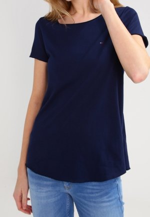 Kvinde iført en ensfarvet mørkeblå t-shirt med korte ærmer og lyseblå jeans, med den ene hånd, der rører ved hendes hår.