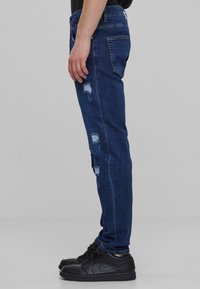 Mørkeblå denim jeans med tettsittende passform, som har slitte lapper og fem lommer. Brukt med svarte sneakers.