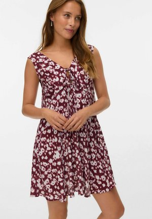 Vrouw in een mouwloos maroon en wit bloemenjurk met een V-hals en een voorknoopdetail, staand met haar handen samengevouwen.